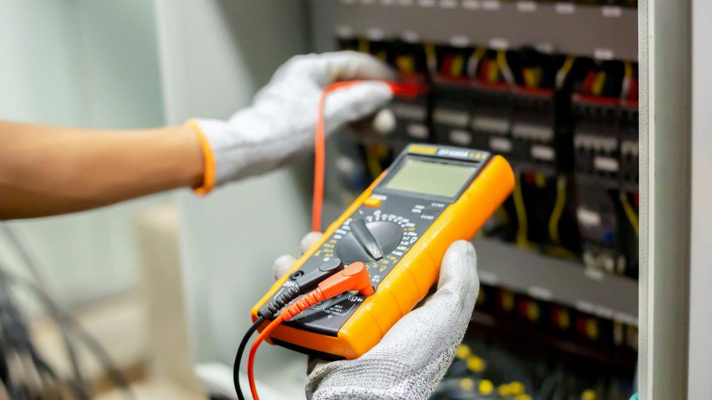 A person wearing gloves and holding a multimeter in front of an electrical panel